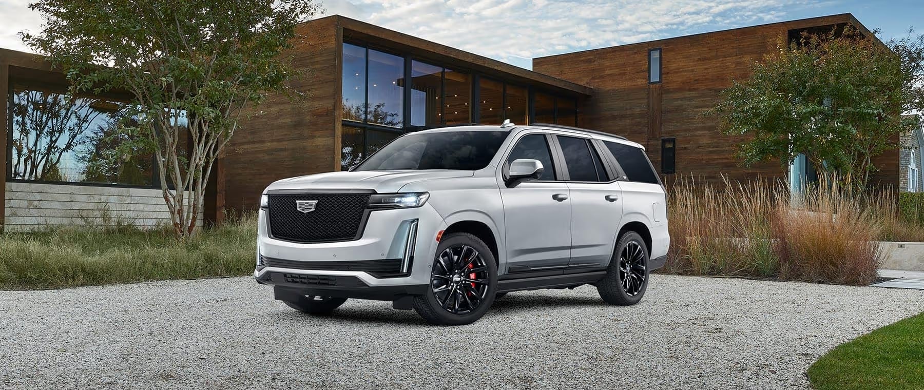 Luxury white Cadillac SUV parked in a modern residential setting with a wooden house in the background, showcasing sleek design and black alloy wheels.