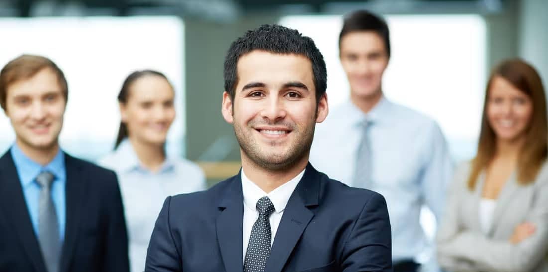 Group of confident business professionals posing together in an office setting, with a focus on a smiling man in a suit, highlighting teamwork and corporate success.