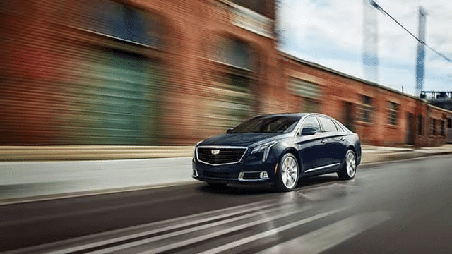 A sleek blue Cadillac XTS sedan speeding down an urban street with brick buildings in the background, showcasing its modern design and performance capabilities.