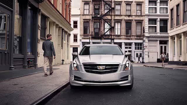 A stylish silver Cadillac sedan parked on a city street, with a casually dressed man walking away in the background, showcasing an urban environment with classic architecture.