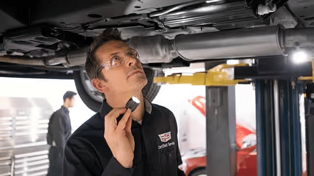 Automobile technician inspecting the underside of a vehicle while using a flashlight, with another technician visible in the background, in a well-equipped auto repair shop.