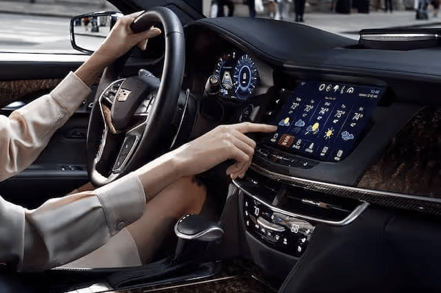Person using a touchscreen display in a luxury car, showcasing navigation and weather features, with a close-up of hands on the steering wheel and console controls.