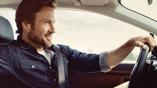 Smiling man with a beard driving a car, showcasing an enjoyable journey in a modern vehicle. Natural light highlights his relaxed demeanor and casual attire.
