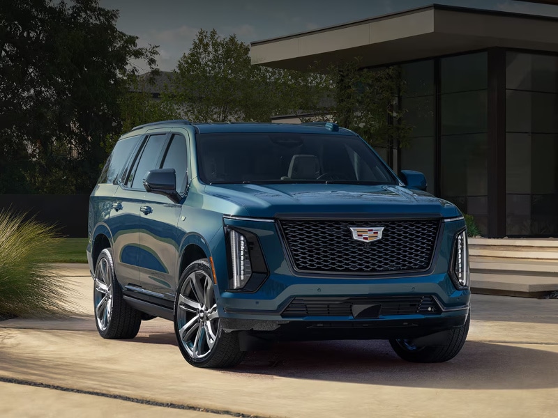 2025 Cadillac Escalade SUV in blue parked in a modern outdoor setting, showcasing its bold front grille, sleek design, and luxury features.