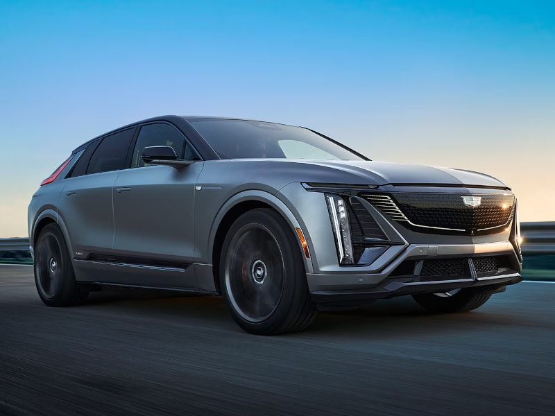 Sleek silver Cadillac electric SUV in motion, showcasing aerodynamic design and modern features against a clear blue sky. Perfect representation of luxury performance vehicles.