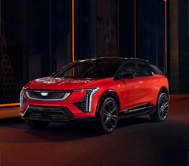 Red electric SUV parked in a modern urban setting, showcasing sleek design and futuristic features, with LED headlights and a stylish black roof.
