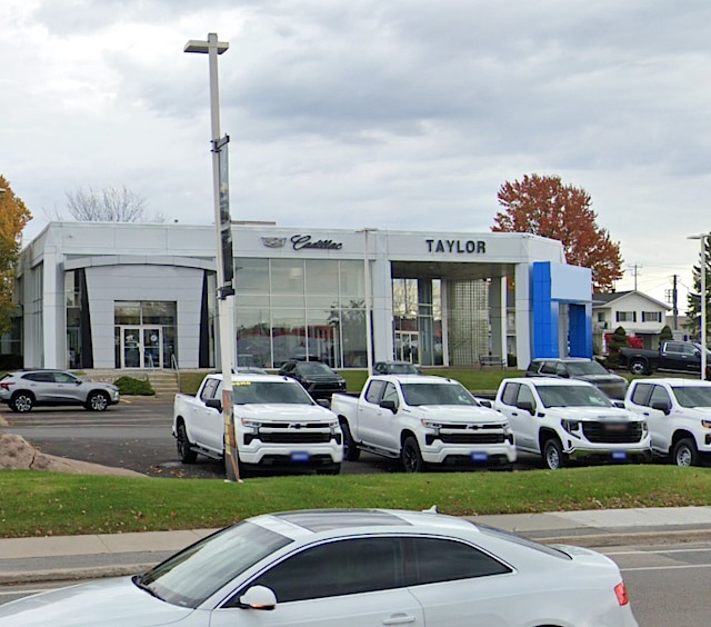 Cadillac dealership exterior with large glass entrance and the name "Taylor" prominently displayed, featuring several white vehicles parked out front on a cloudy day.