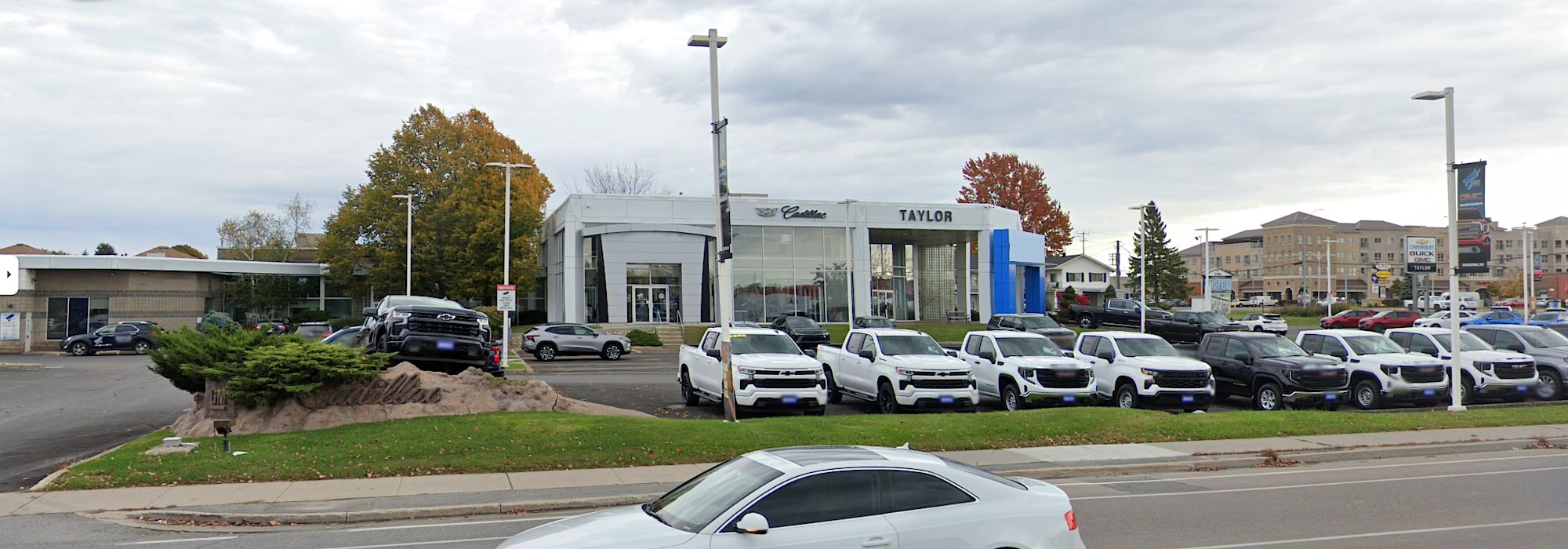 Exterior view of Taylor Cadillac dealership featuring a variety of vehicles, including pickup trucks and SUVs, displayed in the lot. The modern building showcases the dealership's branding, surrounded by trees and a cloudy sky. This image highlights the auto sales environment and available inventory.