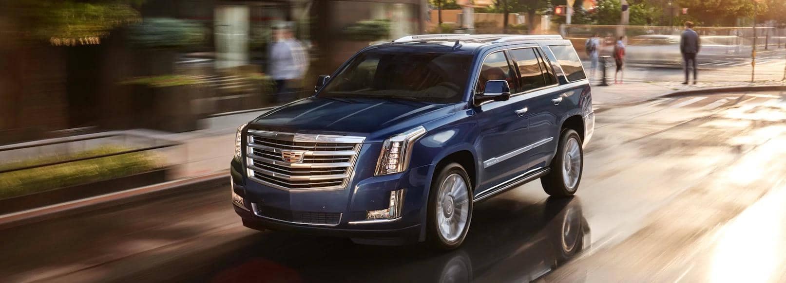A blue Cadillac Escalade driving dynamically down a city street, showcasing its sleek design and luxury features against a blurred urban backdrop.