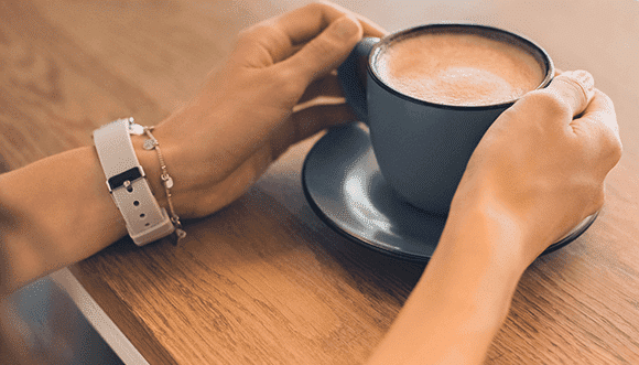 Person holding a blue coffee cup on a saucer, with a wooden table in the background, showcasing a casual coffee moment.