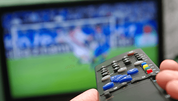 Person holding a TV remote with a blurred image of a soccer game on the screen in the background, illustrating the experience of watching sports at home.