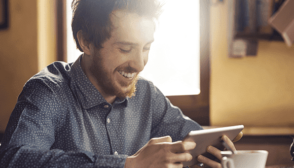 Smiling young man wearing a polka-dot shirt, using a tablet in a cozy café environment, with sunlight streaming through the window, enjoying his time.
