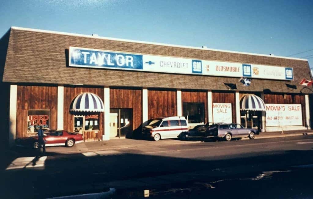Vintage car dealership building exterior with signs for Taylor Chevrolet, Oldsmobile, and Cadillac. Displays a "Used Cars" sign and a "Moving Sale" banner. Several parked cars are visible, including a red sports car and a white van.