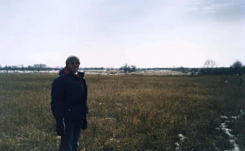 Person standing in a wide, grassy field with sparse trees in the background, under a clear sky during winter, showcasing a peaceful rural landscape.