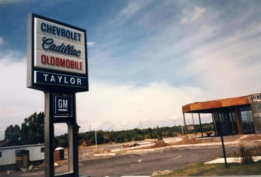 Historic Chevrolet, Cadillac, and Oldsmobile dealership sign with Taylor branding, set against a backdrop of a construction site and clear blue sky. The image captures the automotive heritage and changing landscape of the area.