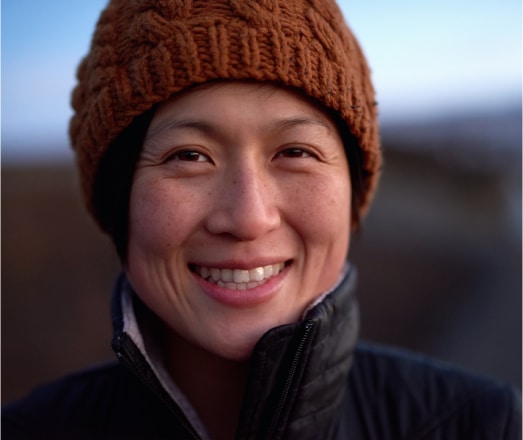 Smiling woman wearing a knitted brown beanie and a black jacket, with a softly blurred outdoor background, showcasing a warm, friendly expression.