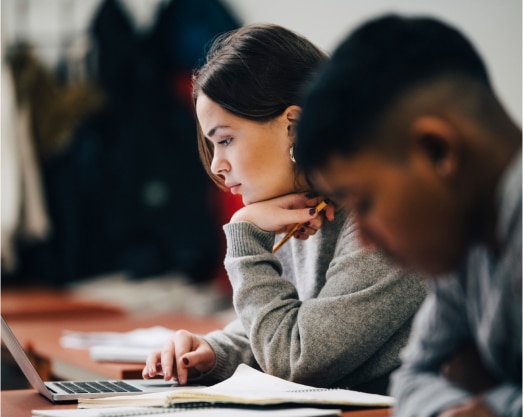 Focused students studying together, one female with a laptop and notebook, and one male in the background, in a collaborative learning environment.