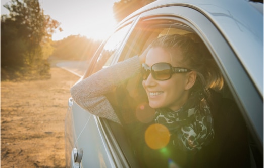 A woman enjoying a scenic drive with her arm resting outside a car window, wearing sunglasses and a scarf, basking in the sunlight on a winding road surrounded by nature.