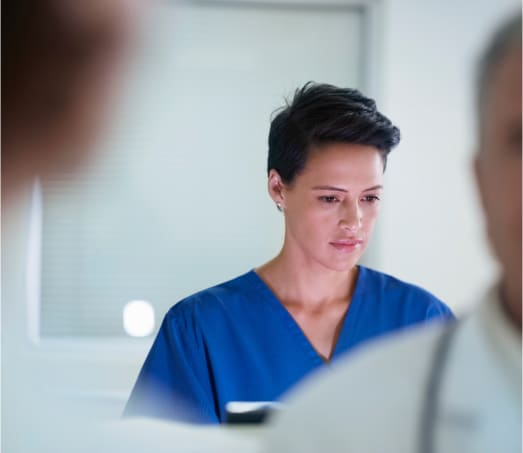 A focused medical professional in a blue scrub top, attentively analyzing information in a clinical setting, with two blurred colleagues in the foreground.