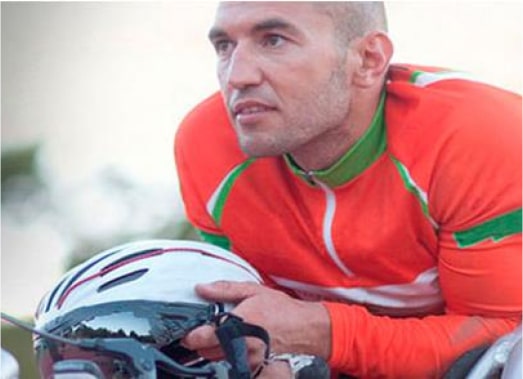 Athlete in a bright orange cycling jersey holding a white helmet, appearing focused and determined, ready for a cycling competition or training session.