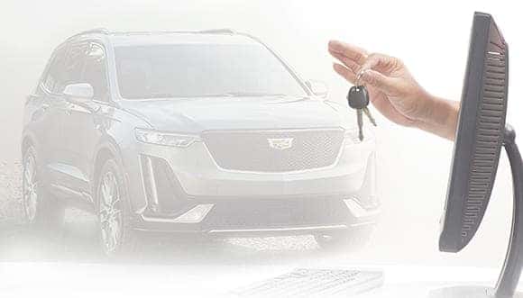Person holding car keys in front of a computer monitor, with a Cadillac SUV in the background, symbolizing online car buying or dealership services.