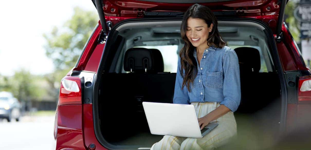 Woman sitting in the trunk of a red SUV, working on a laptop outdoors, smiling and enjoying a casual workspace in a sunny environment.