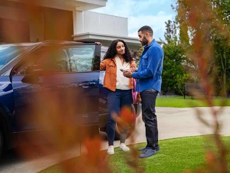 A man and woman discuss details while standing by a blue car in a residential driveway, surrounded by greenery. The woman holds a bag and appears engaged, while the man checks information on his phone.