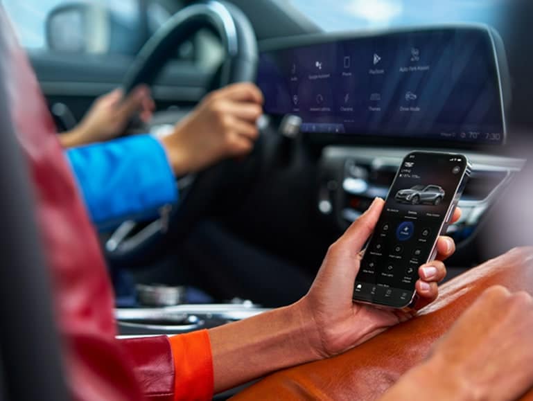 A person in a colorful jacket uses a smartphone to control car features while seated in a luxury vehicle, showcasing modern technology and connectivity in automotive design.