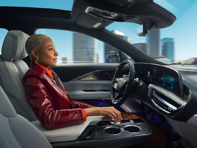 A woman with short blonde hair sits in the driver's seat of a modern car, showcasing a sleek interior with high-tech dashboard displays and a scenic city view through the window. The woman wears a stylish red leather jacket and appears focused on driving.